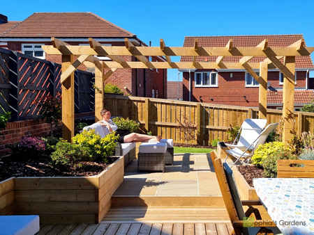 A before image showing a sloping patio with furniture covered up with a dark grey cover. A dark grey, tall fence is situated at the border of the garden, with a neighbours hours overlooking the garden. The after image shows bright blue skies with a lady lounging on a new stone patio with timber raised beds around the edges. The rear fence is now a more natural brown and a wooden pergola shelters the patio from the overlooking neighbours. Click image for more details.