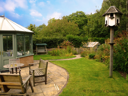 A lawn sweeps around a soft green half-brick conservatory leading to a swing seat next to a pond. There's a patio in the foreground, with seating. Click image for more details.