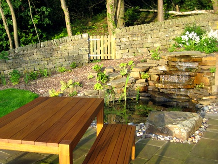 A cedar table & bench seat are placed in front of a natural stone waterfall. Behind is a bark path and low stone wall with a wooden gate leading to the woodland to the rear. Click image for more details.